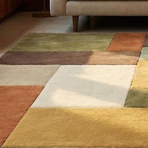 A contemporary wool carpet featuring a color-block patchwork design in soft, muted earth tones. The rectangular rug is centered between sofas, offering comfort underfoot and visual balance to the room. Handmade craftsmanship is visible in the dense pile and clean geometric edges.