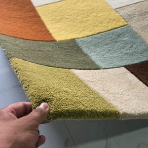 May include: A rectangular area rug featuring a geometric pattern of multi-colored blocks in shades of olive green, mustard yellow, burnt orange, light blue, cream, and dark brown. The rug has a thick, tufted pile texture and a soft, plush surface.
