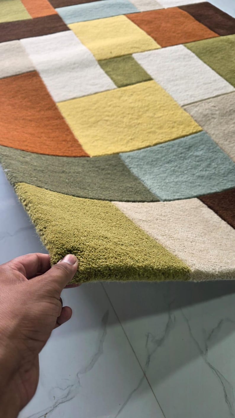 May include: A rectangular area rug featuring a geometric pattern of multi-colored blocks in shades of olive green, mustard yellow, burnt orange, light blue, cream, and dark brown. The rug has a thick, tufted pile texture and a soft, plush surface.