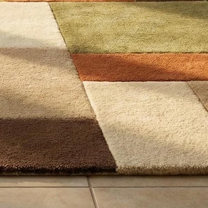 Modern geometric wool rug handcrafted with high-quality natural wool. The patchwork pattern includes warm shades of green, yellow, rust, brown, and cream, forming a balanced and stylish composition. Displayed in a contemporary living room, the rug anchors the seating area and enhances the room’s cozy yet sophisticated atmosphere.