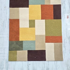 May include: A rectangular area rug featuring a geometric pattern of interlocking blocks in shades of olive green, burnt orange, dark brown, mustard yellow, sage green, and cream. The rug has a textured surface and a small tag on the right side.