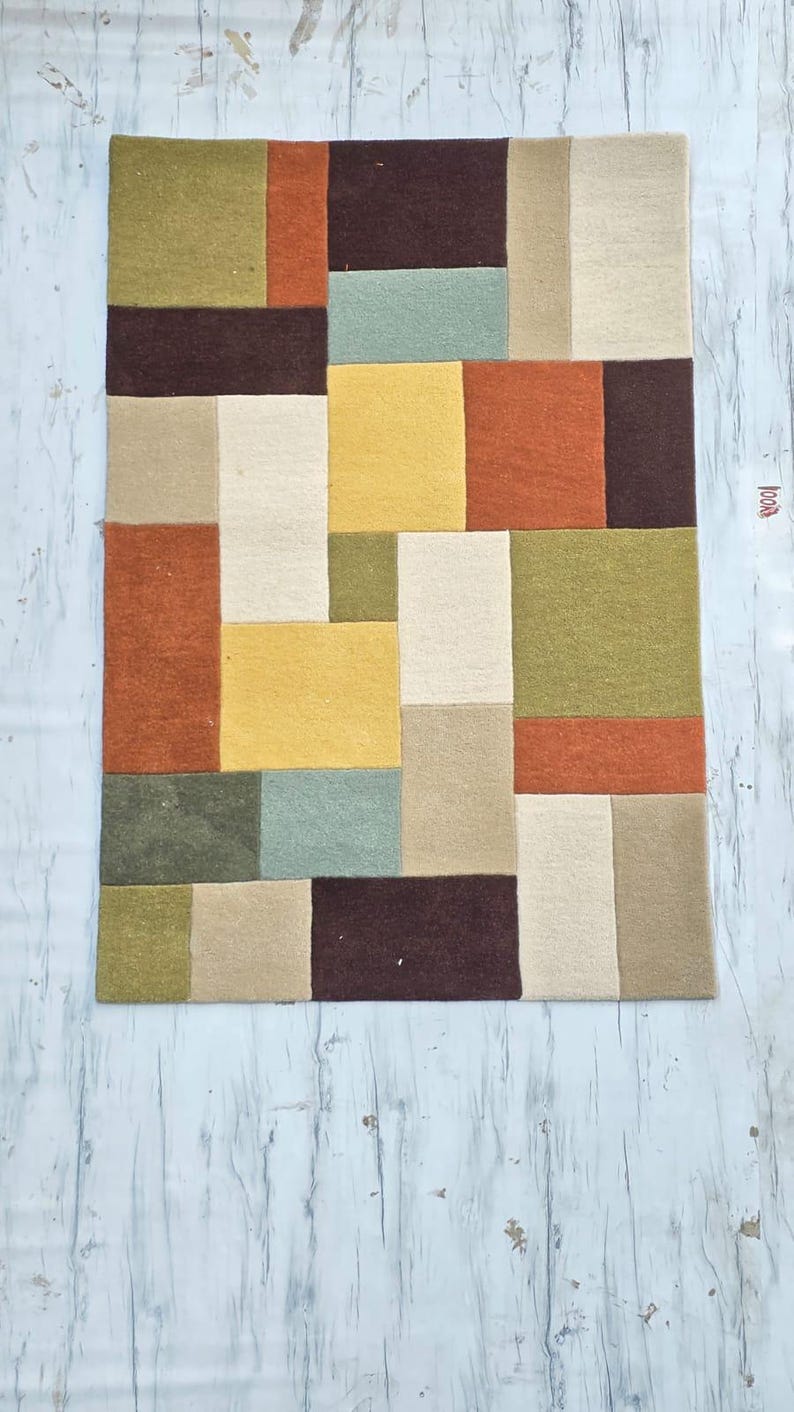May include: A rectangular area rug featuring a geometric pattern of interlocking blocks in shades of olive green, burnt orange, dark brown, mustard yellow, sage green, and cream. The rug has a textured surface and a small tag on the right side.