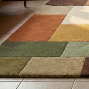 A luxury hand-tufted wool rug featuring a geometric patchwork design inspired by mid-century modern interiors. The rug’s earthy colors blend harmoniously with beige seating, wooden side tables, and natural sunlight. Plush wool fibers create a soft and comfortable surface, while the structured rectangular layout adds visual interest and depth to the living space. Perfect for modern home decor.