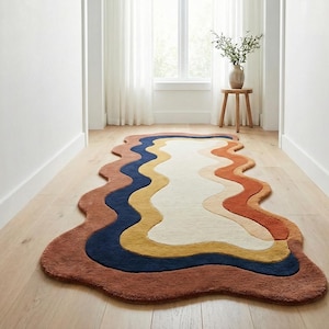 May include: A colorful, wavy-shaped rug with a unique design. The rug features a brown border, with layers of navy blue, yellow, orange, and cream colors. It is placed on a light wood floor in a hallway.