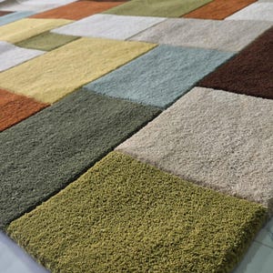 May include: A rectangular area rug featuring a geometric grid pattern composed of plush, textured blocks in various colors including burnt orange, olive green, light yellow, soft blue, dark brown, cream, and sage green, creating a multi-colored surface.