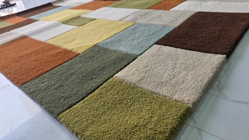 May include: A rectangular area rug featuring a geometric grid pattern composed of plush, textured blocks in various colors including burnt orange, olive green, light yellow, soft blue, dark brown, cream, and sage green, creating a multi-colored surface.