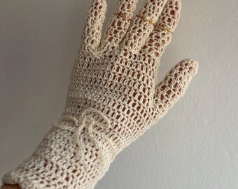 May include: An off-white crochet glove featuring an intricate openwork mesh pattern throughout the hand and fingers. The wrist area includes a decorative tied bow detail and a scalloped edge finish, crafted with fine yarn in a neutral cream tone.
