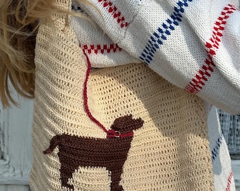 Op de afbeelding: Een beige gehaakte tas met een donkerbruin honden-silhouet als applicatie op de voorzijde. De hond draagt een rode halsband en zit vast aan een dunne rode gehaakte riem die omhoog loopt naar de bovenkant van het handvat van de tas.