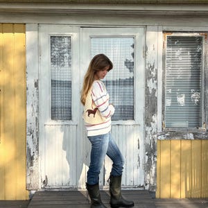 May include: A cream-colored knit sweater featuring horizontal red and blue stripes and a brown dachshund graphic, paired with blue denim jeans and dark green rubber boots with lace-up detailing at the back of the calf.