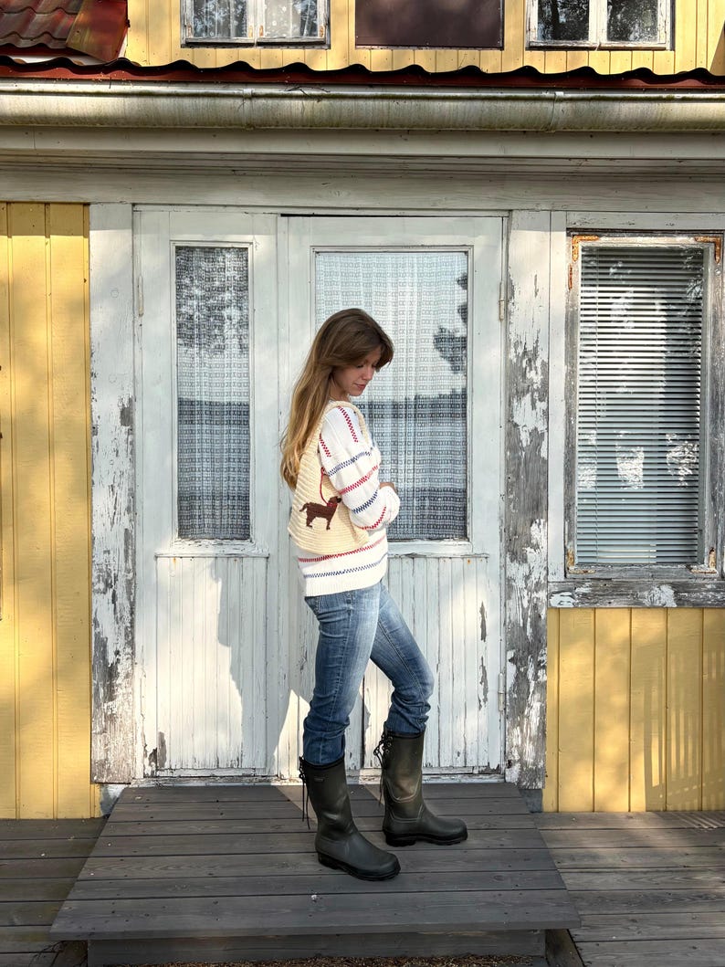 May include: A cream-colored knit sweater featuring horizontal red and blue stripes and a brown dachshund graphic, paired with blue denim jeans and dark green rubber boots with lace-up detailing at the back of the calf.