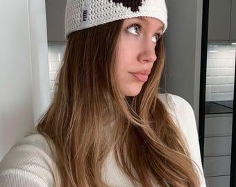 Op de afbeelding: Een witte gehaakte beanie met een donkerbruin hartontwerp. De hoed heeft een klein label met het woord "PERSE". De persoon draagt een witte coltrui. De beanie is een handgemaakt item.