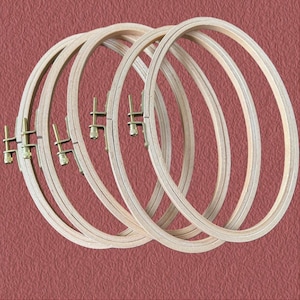 May include: A set of wooden embroidery hoops, overlapping each other. The hoops are made of light-coloured wood and have a visible grain. Each hoop features a brass-toned screw mechanism. The background is a textured, reddish-brown.