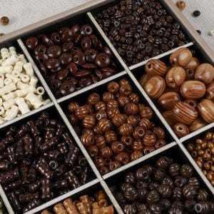 Op de afbeelding: Een houten sorteerdoos met meerdere vakken gevuld met een assortiment houten kralen in verschillende vormen, maten en bruintinten, waaronder cilindrische, ronde en ovale kralen in crème, lichtbruin en donkerbruin.