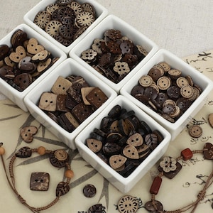 May include: A collection of dark brown coconut shell buttons in various shapes including hearts, squares, flowers, and leaves, organized into five white square ceramic bowls, with two braided cord bracelets featuring button accents.