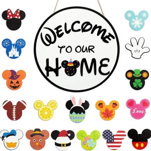 May include: A round white wooden sign with black text reading WELCOME TO OUR HOME, featuring a mouse-ear icon for the letter O. It is surrounded by 18 interchangeable seasonal and themed mouse-ear shaped wooden icons in various colours.