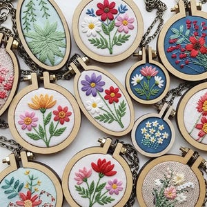May include: A collection of small embroidery hoop necklaces featuring various floral designs stitched onto fabric backgrounds. Each piece is framed in a circular or oval wooden hoop with a metal screw top and attached to a dark metal chain.
