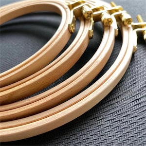 May include: Close-up of several wooden embroidery hoops with brass hardware. The hoops are light brown and arranged in a slightly overlapping pattern. The brass hardware is visible at the top, and the background is a textured gray fabric.