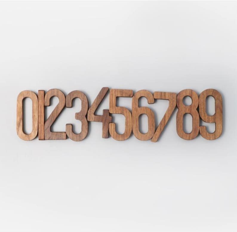 May include: Wooden numbers 0 through 9 in a warm brown tone. The numbers are cut from wood and arranged horizontally against a white background. The numbers are in a sans-serif font.