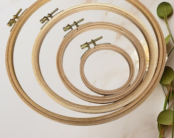 May include: Four wooden embroidery hoops of varying sizes are arranged in a spiral. Each hoop has a brass-colored tightening screw. The hoops are made of light-colored wood and are set against a neutral background, suggesting a craft or DIY theme.