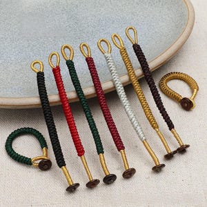 May include: A collection of seven braided cord toggles in black, red, dark green, maroon, silver, gold, and dark brown, each featuring a small loop at one end and a dark circular button fastener at the other, arranged in a row.