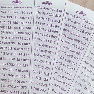 May include: Three white rectangular sheets featuring a grid of purple DMC brand labels. Each label contains a unique numerical or text-based color code for embroidery thread organization. The sheets are arranged in a slight overlap, displaying rows of numbers.