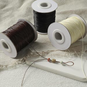 May include: Three spools of cord in dark brown, black, and cream colours, each wound around a white plastic reel. A single dark brown cord extends from the brown spool, threaded through three small, multi-coloured decorative beads.