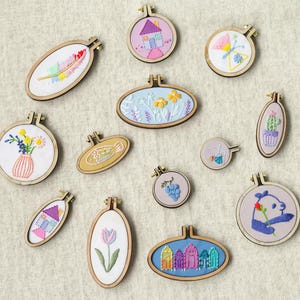 Può includere: Una collezione di piccoli telai da ricamo in legno con vari disegni cuciti, tra cui una casa, una farfalla, fiori in un vaso, un pesce, un cactus, uva, un tulipano, un panda che tiene un fiore e colorate cabine da spiaggia su tessuto.