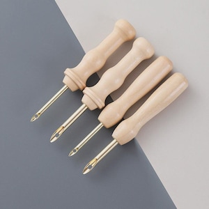 May include: Four rug hooking tools. Each tool has a light-coloured wooden handle and a brass needle. The needles have a small opening near the tip. The tools are arranged diagonally against a grey and white background.