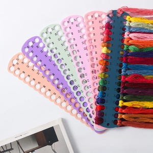 Puede incluir: Una colección de hilos de bordar coloridos y organizadores de hilo. Los organizadores son de varios colores pastel, cada uno con agujeros numerados para la colocación del hilo. El hilo es un arcoíris de tonos.