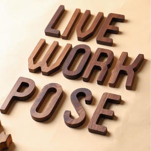 May include: Wooden letters spelling out "LIVE WORK POSE" in a warm brown tone. The letters are thick and three-dimensional, with a natural wood grain visible. The letters are arranged on a light tan surface.