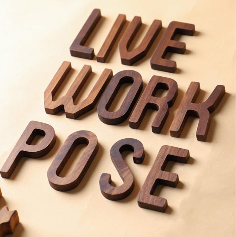May include: Wooden letters spelling out "LIVE WORK POSE" in a warm brown tone. The letters are thick and three-dimensional, with a natural wood grain visible. The letters are arranged on a light tan surface.