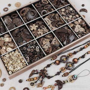 May include: A rectangular organiser box filled with assorted brown and tan coconut shell buttons in various shapes, including circles, triangles, and stars. Beside the box are three black cord necklaces featuring decorative wooden beads and charms.