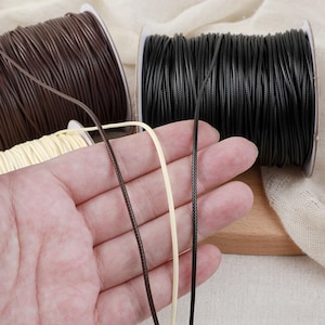 Puede incluir: Tres carretes de cordón en colores marrón oscuro, negro y crema. Los cordones son finos, redondos y presentan una textura trenzada o tejida. Cada carrete está enrollado con una longitud considerable de cordón, y se sostienen hebras individuales.