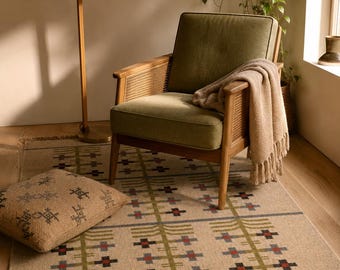May include: A rectangular area rug featuring a beige base with a repeating grid pattern of olive green lines and small cross motifs in red, blue, and black. The rug has a fringed edge and is displayed with a matching square throw pillow.