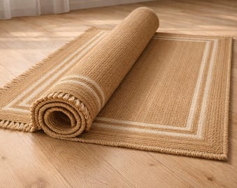 May include: A rolled-up rectangular rug with a fringed edge and a light brown color. The rug has a lighter border and is partially unrolled, revealing the texture and design. The rug is on a wooden floor.