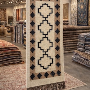 May include: A rectangular beige rug with a geometric pattern of black and brown shapes. The rug has a brown border and fringe at the bottom. The rug is hanging in a rug store, with other rugs visible in the background.