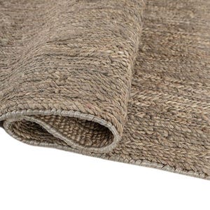 May include: Close-up of a rolled-up, neutral-toned rug. The rug has a textured, woven appearance with visible fibers and a natural, earthy color palette. The edge of the rug is visible, showing a finished border.