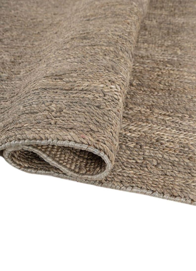 May include: Close-up of a rolled-up, neutral-toned rug. The rug has a textured, woven appearance with visible fibers and a natural, earthy color palette. The edge of the rug is visible, showing a finished border.