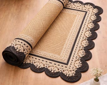 May include: Rolled-up beige and black area rug with a scalloped edge. The rug has a woven texture and a decorative border. The rolled section reveals the underside, which is black. The rug is partially unrolled on a wooden floor.