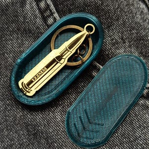 Op de afbeelding: Een goudkleurige metalen sleutelhanger in de vorm van een kogel met de tekst 122MM gegraveerd op de zijkant, liggend in een ovaalvormig, blauwgroen 3D-geprint doosje met een bijpassend deksel voorzien van drie ingedrukte chevron-patronen.