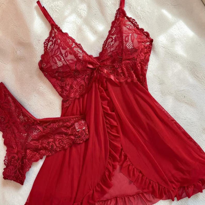 A vibrant wine red lace lingerie set laid flat against a soft, textured background features a sheer nightdress with a deep v-neckline, delicate floral lace cups, thin shoulder straps, and a front bow accent alongside matching lace panties.