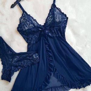 A vibrant navy blue lace lingerie set laid flat against a soft, textured background features a sheer nightdress with a deep v-neckline, delicate floral lace cups, thin shoulder straps, and a front bow accent alongside matching lace panties.