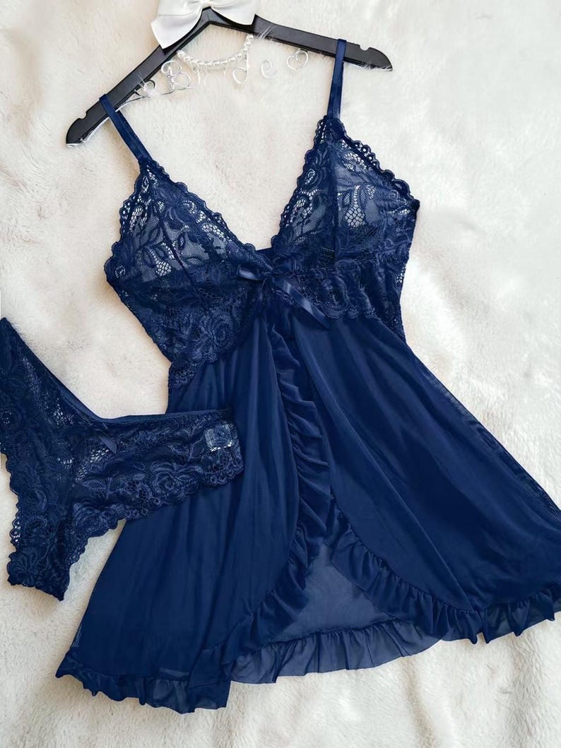 A vibrant navy blue lace lingerie set laid flat against a soft, textured background features a sheer nightdress with a deep v-neckline, delicate floral lace cups, thin shoulder straps, and a front bow accent alongside matching lace panties.