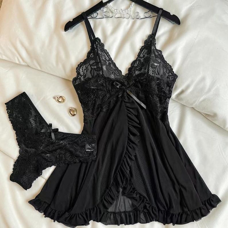 A vibrant black lace pajama set laid flat against a soft, textured background features a sheer nightdress with a deep v-neckline, delicate floral lace cups, thin shoulder straps, and a front bow accent alongside matching lace panties.
