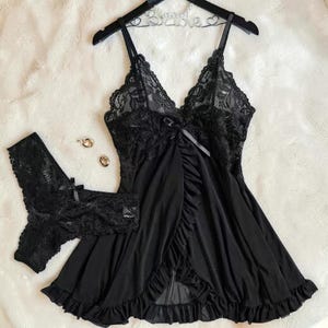 A vibrant black lace lingerie set laid flat against a soft, textured background features a sheer nightdress with a deep v-neckline, delicate floral lace cups, thin shoulder straps, and a front bow accent alongside matching lace panties.