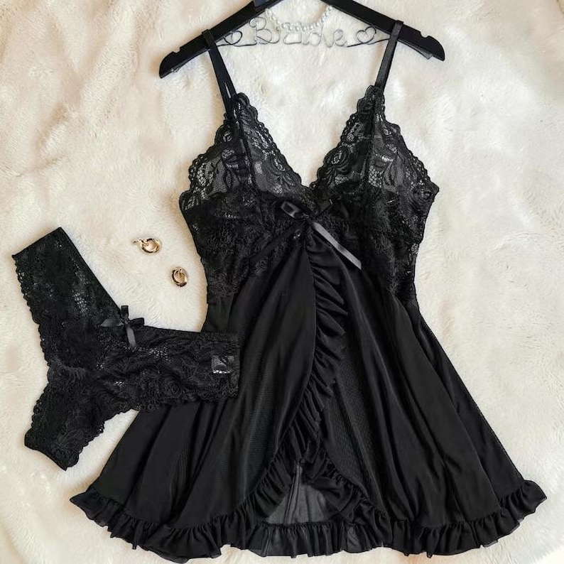 A vibrant black lace lingerie set laid flat against a soft, textured background features a sheer nightdress with a deep v-neckline, delicate floral lace cups, thin shoulder straps, and a front bow accent alongside matching lace panties.