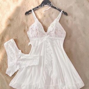 A vibrant white lace pajama set laid flat against a soft, textured background features a sheer nightdress with a deep v-neckline, delicate floral lace cups, thin shoulder straps, and a front bow accent alongside matching lace panties.