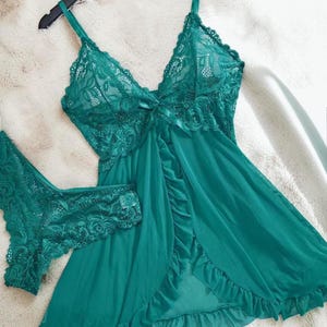 A vibrant peacock green lace lingerie set laid flat against a soft, textured background features a sheer nightdress with a deep v-neckline, delicate floral lace cups, thin shoulder straps, and a front bow accent alongside matching lace panties.