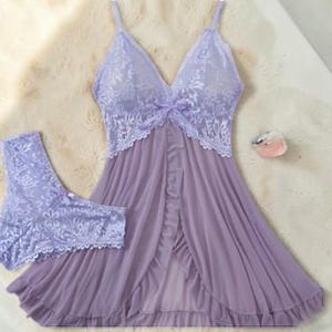 A vibrant purple lace lingerie set laid flat against a soft, textured background features a sheer nightdress with a deep v-neckline, delicate floral lace cups, thin shoulder straps, and a front bow accent alongside matching lace panties.