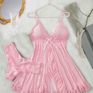 A vibrant pink lace lingerie set laid flat against a soft, textured background features a sheer nightdress with a deep v-neckline, delicate floral lace cups, thin shoulder straps, and a front bow accent alongside matching lace panties.